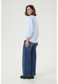 Blouse bleu clair avec des pois texturés, encolure arrondie et manches bouffantes, associée à un jean en denim foncé à jambes larges et des chaussures à plateforme noires.