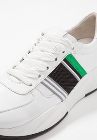 Białe sneakersy z siateczki i materiału skórzanego, z zielonymi i szarymi paskami, okrągłym noskiem i białymi sznurowadłami. Czarna gumowa podeszwa.