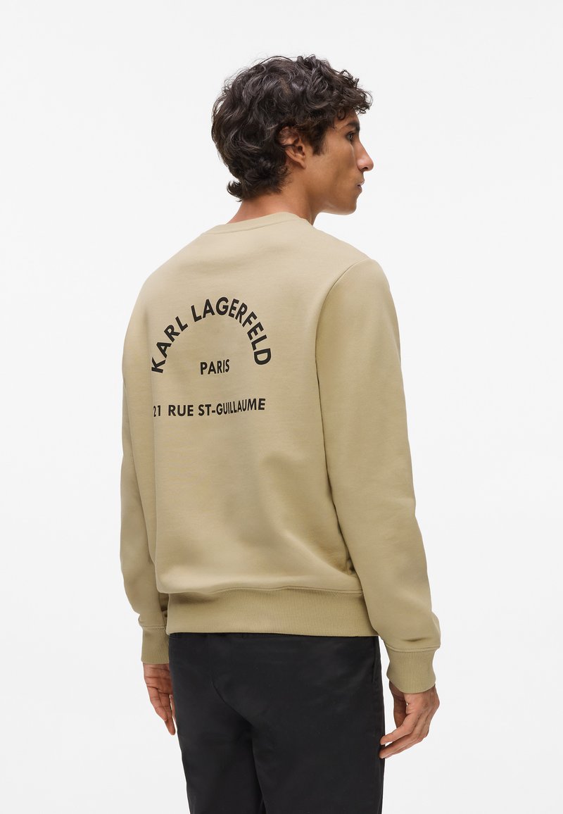 Béžová mikina s rebrovanými manžetami a spodným lemom. Veľký čierny nápis na chrbte znie "KARL LAGERFELD PARIS 21 RUE ST-GUILLAUME." Jednoduchý dizajn, bez vzorov.