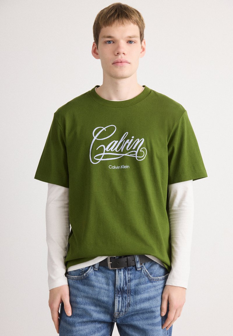 Young man wearing layered green Calvin Klein T-shirt and white long sleeves tucked into blue jeans with black belt, standing against white background.