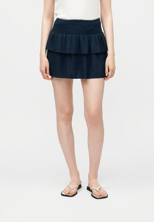 Woman wearing a navy blue tiered mini skirt, white sleeveless top, and beige square-toe sandals, standing against a white background.