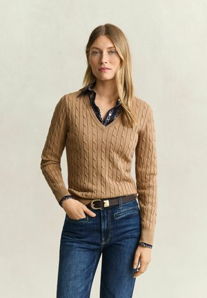 Femme aux longs cheveux blonds portant un pull beige torsadé à col en V par-dessus une chemise sombre à motifs et un jean bleu avec une ceinture marron.