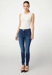 Svijetlosmeđi top bez rukava uparen s tamnoplavim skinny trapericama. Traperice imaju strukirani dizajn i blago izblijedjeli izgled.