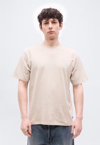 Νεαρός άνδρας με σγουρά σκούρα μαλλιά, φορώντας μπεζ t-shirt Levi's και μπλε τζιν, στέκεται μπροστά σε ένα απλό φωτεινό φόντο.