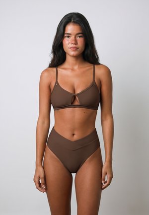 Jonge vrouw met lang donker haar die een bruine bikini met uitgesneden top en hoogsluitende broek draagt, staande tegen een effen lichte achtergrond.