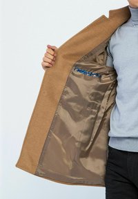 Brauner Wollmantel mit glattem, beigem Futter, ausgestattet mit einer kleinen Innentasche mit blauer Musterung. Klassisches Design mit einer klaren, maßgeschneiderten Passform.