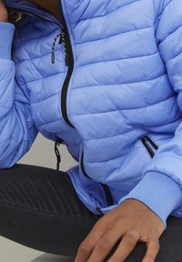 Persona con una chaqueta acolchada azul clara y cremalleras negras, y pantalones oscuros, sentado con una mano descansando sobre su rodilla.