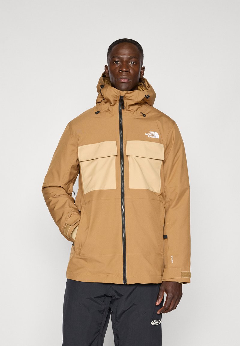 Giacca impermeabile marrone con tasche sul petto beige, cappuccio regolabile e zip nera. Tessuto testurizzato con un design funzionale per l'uso all'aperto.