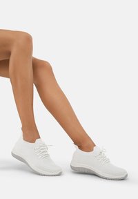 Baskets en tricot blanches avec un dessus texturé, des lacets élastiques et une semelle en caoutchouc grise contrastante avec un design nervuré.