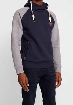 Mann trägt einen marineblauen und grauen Kapuzenpullover mit weißen Kordeln und dunklen Jeans, steht vor einem schlichten hellen Hintergrund.