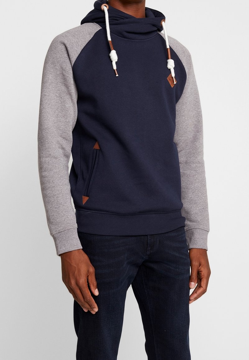 Homme portant un sweat à capuche bleu marine et gris avec des cordons blancs et un jean foncé, debout devant un fond clair uni.