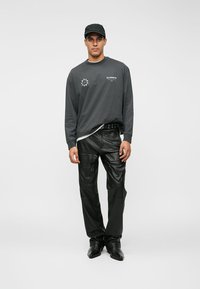 Siva majica dugih rukava s logom, uparenja s crnim kožnim hlačama. Model nosi crnu kapu i tamne cipele, stoji uz jednostavnu pozadinu.