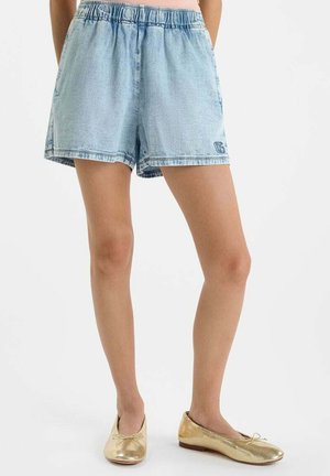 Lichtblauwe denim shorts met elastische tailleband gedragen door een persoon die staat, gecombineerd met gouden ballerina's op een witte achtergrond.