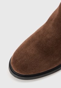 Bottine en suède marron avec un bout arrondi et une semelle en caoutchouc noire. Texture lisse avec des variations subtiles de couleur. Pas de détails métalliques visibles.