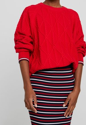 Personne portant un pull rouge à torsades avec les manches retroussées et une jupe côtelée à rayures horizontales rouges, blanches et noires.