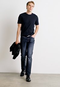 Mørkeblå bomulds T-shirt parret med mørk denim jeans og sorte lædersko. Modellen holder en sort jakke og står foran en lys baggrund.