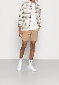 Károvaná košile v bílé, hnědé a černé barvě přes bílém tričku. Khaki cargo šortky. Bílé tenisky a ponožky. Hladké materiály, uvolněný střih.