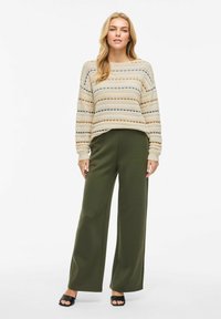 Beige gebreide trui met patronen in verschillende kleuren, gecombineerd met donkergroene wijde pantalons en zwarte hoge sandalen.