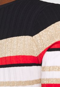 Stickad plagg med svarta, vita, röda och gyllene ränder; texturerade horisontella mönster med glittrande metalliska accenter.