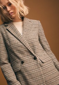 Blazer a quadri in tonalità beige e marrone, caratterizzato da un design doppiopetto, rever a lancia e due tasche frontali.