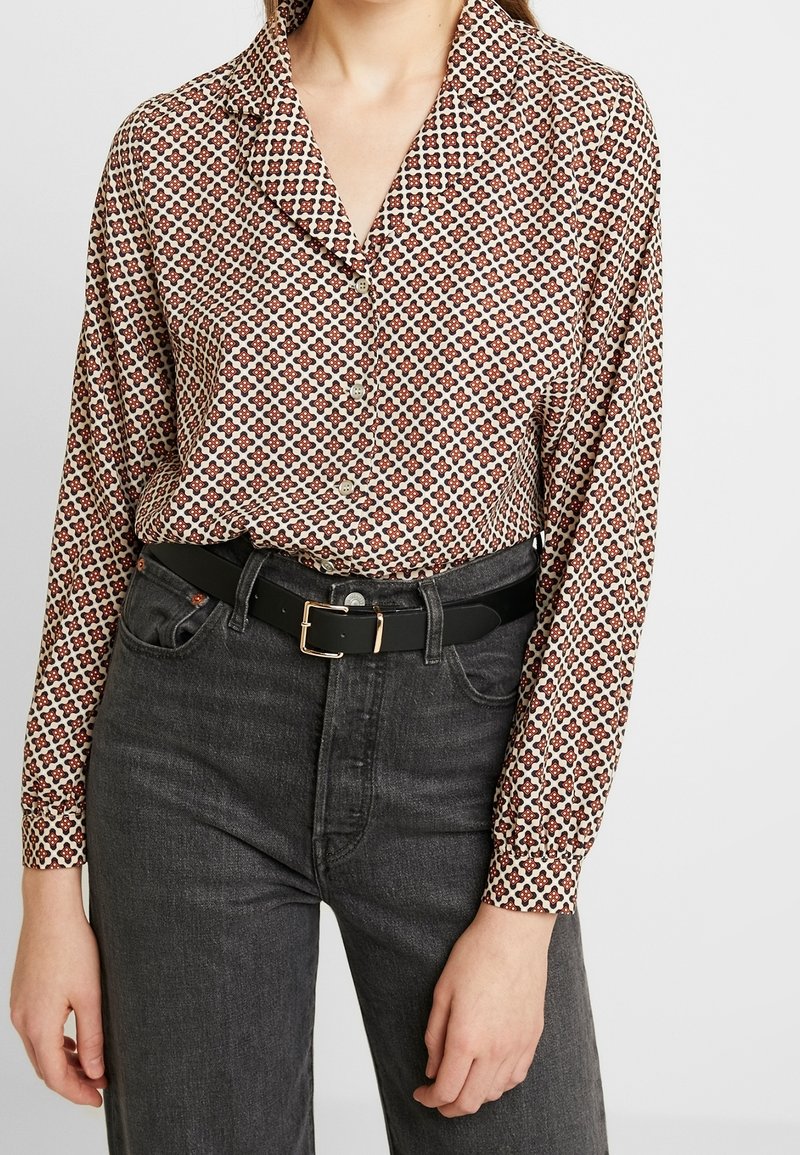 Blusa a maniche lunghe color crema con un motivo floreale rosso e marrone, con colletto e bottoni, abbinata a jeans grigi scuri a vita alta e una cintura nera.