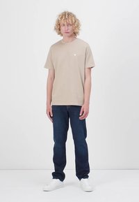 T-shirt en coton beige à manches courtes, avec un petit logo triangulaire. Porté avec un jean en denim noir et des baskets blanches.