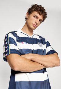 En ung mand med krøllet hår står med armene over kors, iført en navy og hvid stribet sports jersey med Champion logoer på ærmerne og brystet.