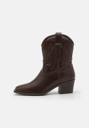 Botte western en cuir brun foncé mi-mollet avec bout pointu, coutures décoratives, languette de tirage et talon bloc empilé sur fond blanc.