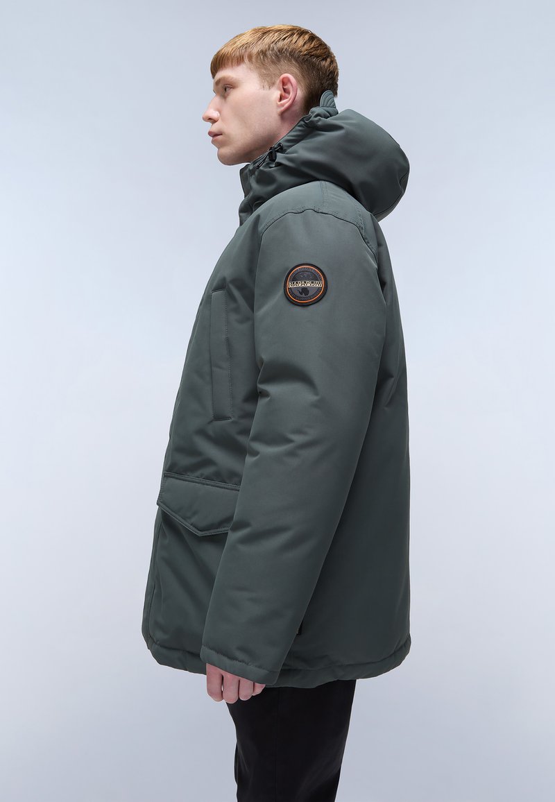 Veste isolante vert foncé avec col montant et capuche, dotée d'une poche poitrine et d'un patch rond sur la manche gauche. Texture lisse.