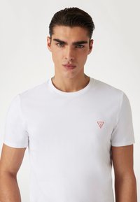 Hvid kortærmet T-shirt lavet af bomuld, med et lille rødt trekantet logo på venstre bryst. Rund halsudskæring, ensartet struktur.