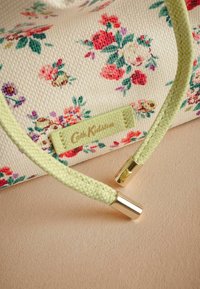 Blomsterdesign på en cremet tekstilpose med grønne vævede håndtag og hardware i guldtoner. Logoetiket i grøn. Glat tekstur.