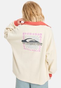 Krémově bílá fleecová bunda s oranžovým límcem, na zadní straně je grafický design v pastelových barvách. Volný střih s dlouhými rukávy.