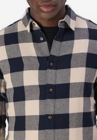 Man in een marineblauwe en beige grote geruite button-up shirt over een zwart T-shirt, zichtbaar van nek tot taille.