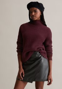 Burgunderfarbener Strickpullover mit hohem Kragen und gerippten Bündchen, über einem schwarzen Leder-Minikilt mit asymmetrischem Saum getragen.