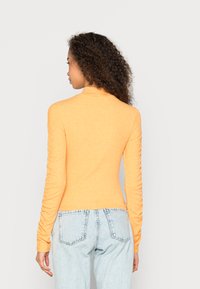 Suéter de manga longa e gola alta em laranja brilhante, com detalhes franzidos nas mangas, combinado com jeans de cintura alta em azul claro.