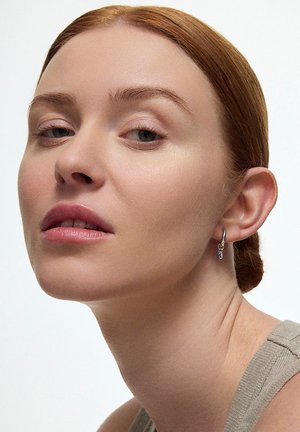 Srebrna kolczyk w kształcie obręczy z małym niebieskim kamieniem akcentującym. Noszony przez modelkę na neutralnym tle, prezentującym design kolczyka.