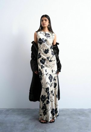 Kvinde iført en lang cremefarvet kjole med sorte blomsterprint, sort pelsfrakke draperet over arme, sorte sandaler og markante øreringe.