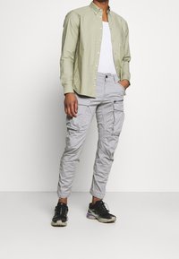 Jasnozielona, zapinana na guziki koszula, biały top na ramiączkach oraz szare spodnie typu cargo z wieloma kieszeniami. Czarne sneakersy z fioletowymi akcentami.