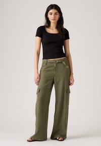 Donna con lunghi capelli scuri che indossa una maglietta nera a maniche corte, pantaloni cargo larghi verde oliva e sandali neri, in piedi di fronte a uno sfondo neutro.