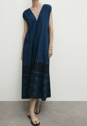 Granatowa sukienka bez rękawów z głębokim dekoltem w serek, z czarnym geometrycznym wzorem u dołu, noszona z beżowymi płaskimi sandałami na szarym tle.