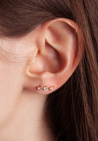 Trzy małe kolczyki typu wkrętki w różnych rozmiarach, z przezroczystymi kryształkami osadzonymi na różowej złotej bazie, przymocowane do płatka ucha.