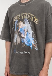 Dunkelgraues, übergroßes T-Shirt aus Baumwolle mit einem grafischen Druck eines Engels in blauem Kleid neben einem Löwen, darunter Text.