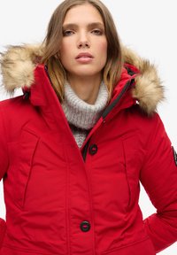 Red padded parka jacket with a faux fur hood, featuring a zip closure, two front pockets, and a high-collar gray knit sweater underneath.