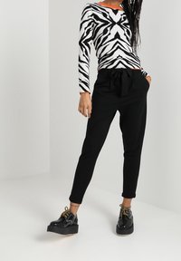 Zwart-wit gemusterd lange mouwen shirt met V-hals, gecombineerd met zwarte slim-fit broek en zwarte plateau schoenen met gouden veters.