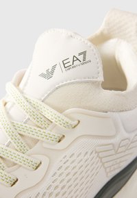 Close-up of cream-colored Emporio Armani EA7 sneaker showing textured fabric, detailed laces, and branded logos on tongue and side.