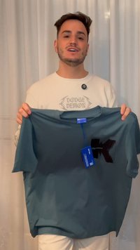 Egy férfi sötétkék pólót tart, rajta címkék, fehér függönyök előtt áll, fehér pulóvert visel, amelyen a "DODGE DEMON" felirat szerepel.