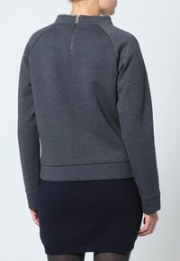 Donna che indossa un top a maniche lunghe grigio scuro con zip sul retro e una gonna fitted blu scuro, in piedi contro uno sfondo chiaro e uniforme.
