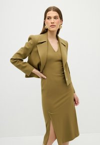 Olijfgroen blazer met gestructureerde schouders, gecombineerd met een aansluitende jurk met V-hals en zijsplit. Gouden knoopaccenten op de blazer.