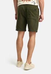 Un shorts vert olive en tissu texturé, avec une poche arrière et une coupe ample, associé à des baskets beiges.