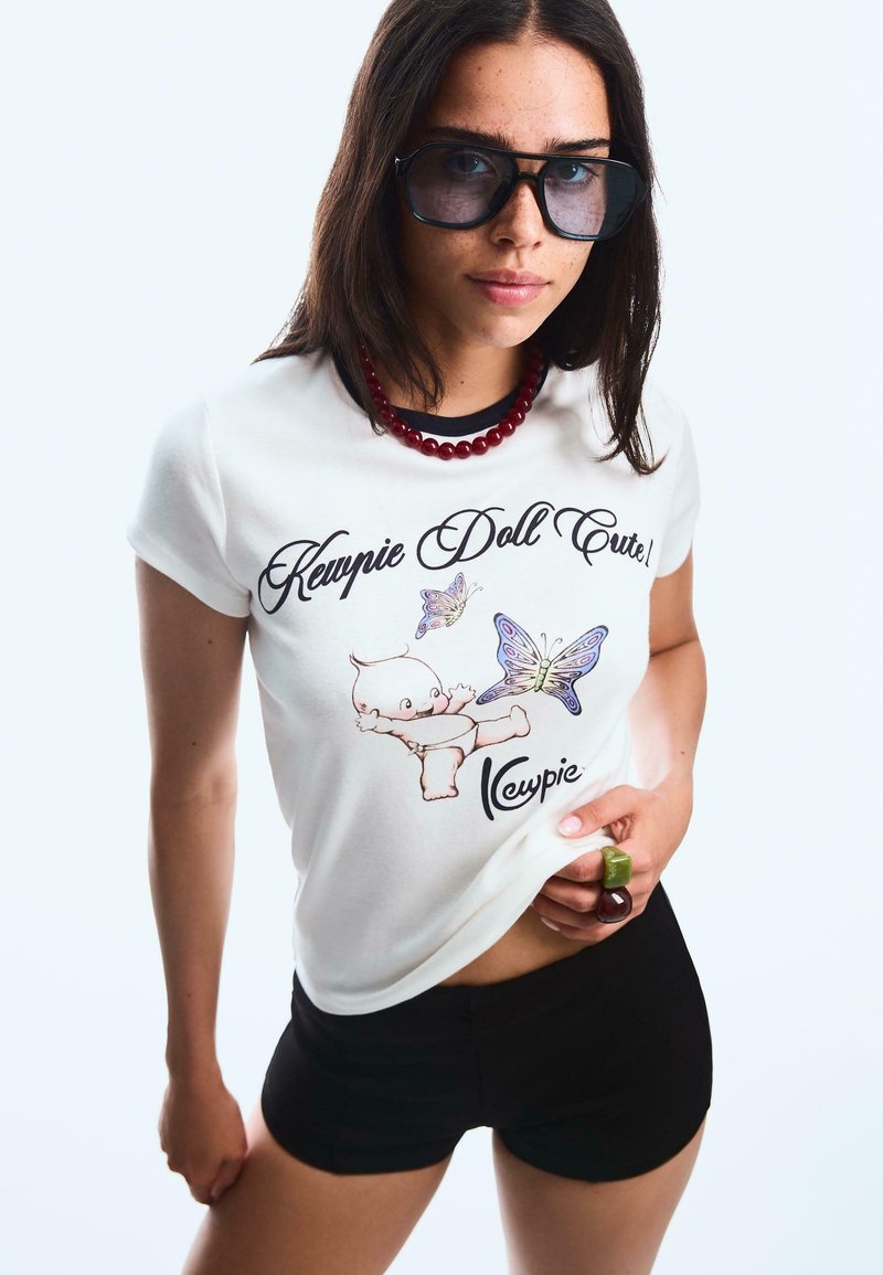 White t-shirt with "Kewpie Doll Cute!" print, featuring a Kewpie doll and butterflies. Black shorts and a red bead necklace add contrast. Large sunglasses.